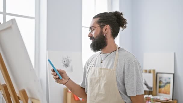 Um jovem barbudo em um avental sorri enquanto usa um smartphone em um estúdio de arte brilhante. - Filmagem, Vídeo