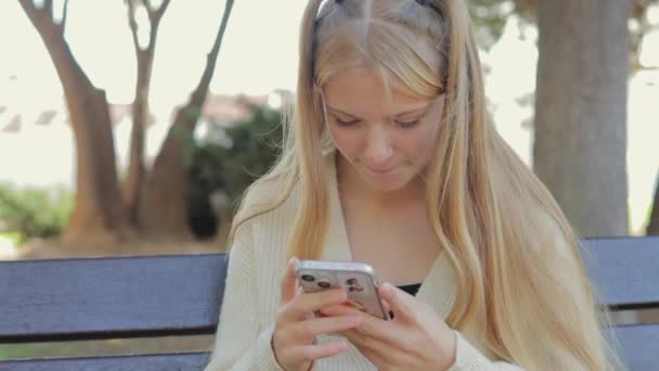 meisje in een wit shirt zit op de bank en maakt gebruik van een smartphone, typen in het park - Video