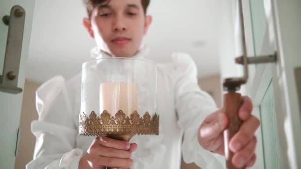 Serene young man in a white ruffled shirt holding a candle holder, gazing out a bright window in a minimalist room during the daylight - Footage, Video