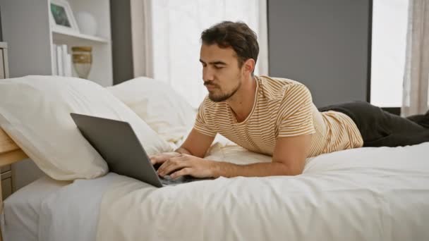 Un joven hispano con barba acostado en una cama trabajando en un portátil en un dormitorio moderno - Metraje, vídeo
