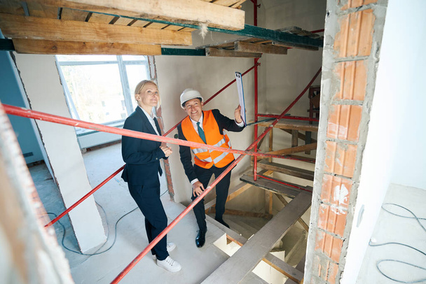 Full length photo of amused businesswoman and smiling construction foreman standing under scaffolding while inspecting unfinished house - Photo, Image