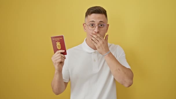 Shocked young hispanic man covers mouth with hand, holding denmark passport. silent surprise, fear of a grave mistake, isolated on yellow background - Footage, Video