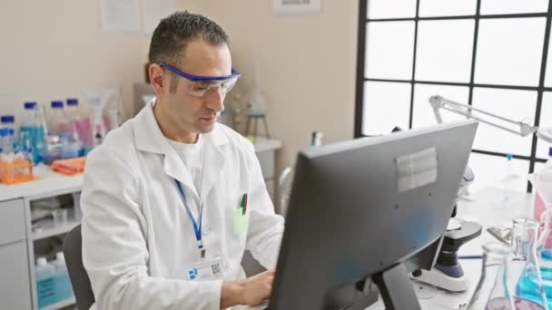 Een professionele Spaanse man in een labjas werkt intensief op een computer in een heldere laboratoriumomgeving. - Video