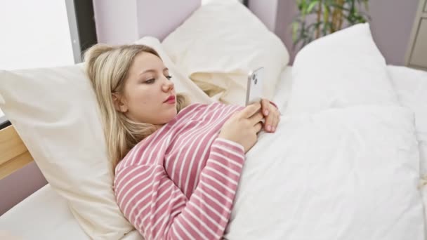 Une femme blonde en chemise rayée se trouve au lit engagée avec son smartphone dans un cadre lumineux de chambre à coucher. - Séquence, vidéo