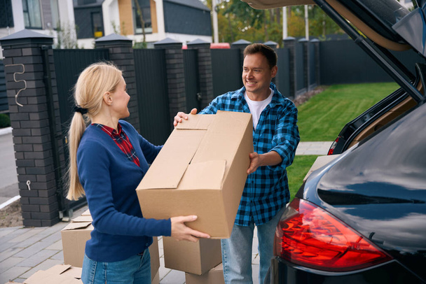 Smiling man unloading cardboard box out of car trunk and giving it to happy woman standing near him - Photo, Image