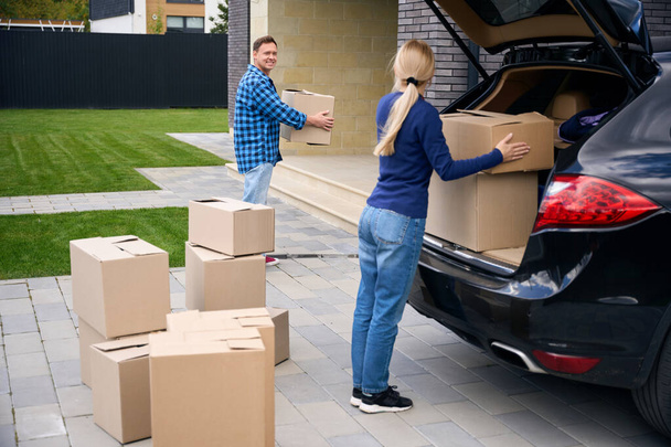 Pełna długość zdjęcie kobiety rozładowującej kartonowe pudełko z samochodu i mężczyzny trzymającego kolejne podczas jazdy do domu - Zdjęcie, obraz