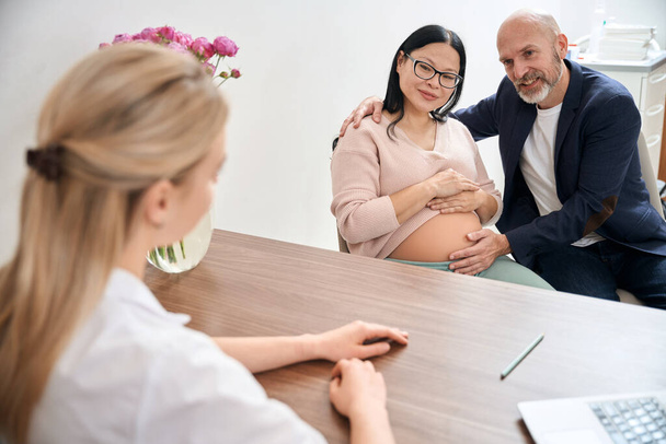 Volwassen familie paar bezoeken verloskundige kantoor om te praten over het derde trimester van de zwangerschap en het proces van bevalling met haar hulp, contract bevalling - Foto, afbeelding
