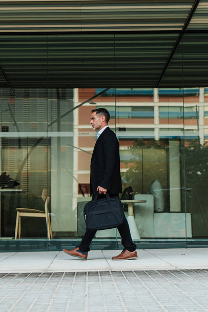Vertical full length portrait of a mature business man walking and holding a laptop bag. Mid adult financial executive male going to the office. Handsome corporate business person with a briefcase - Photo, Image