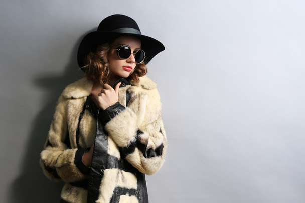 Portrait of beautiful model in fur coat, hat and sunglasses on gray background - Photo, Image