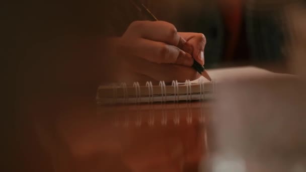 Zbliżenie osoby piszącej w notatniku na drewnianym stole z zamazanym pierwszym planem w godzinach porannych - Materiał filmowy, wideo