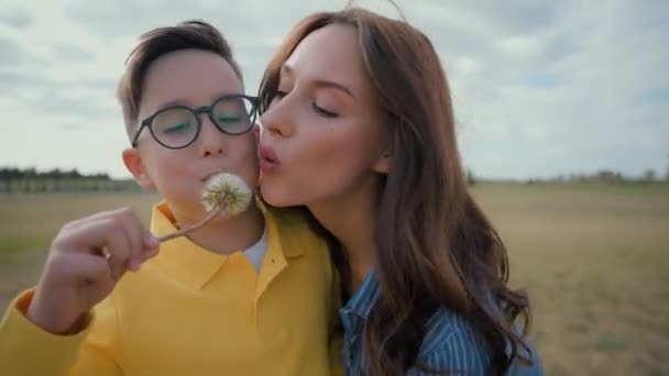 幸せな子供時代のコーカサス家族笑顔の母親の小さな男の子の息子はタンポポの花を吹き飛ばして楽しいリラクゼーション休暇を持っている街の外で自然のフィールドをかわいがって楽しい夏の思い出の親 - 映像、動画