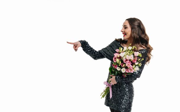 Beautiful young woman in black dress is holding bouquet of flowers and pointing at something on white background. The concept of holiday birthday Valentine's Day. - Foto, Imagen