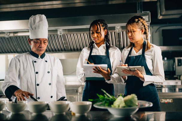 In a restaurant kitchen an Asian senior chef in uniform educates multiracial trainees. Teacher imparts cooking skills emphasizing teamwork learning and note-taking in this professional workshop. - Fotoğraf, Görsel