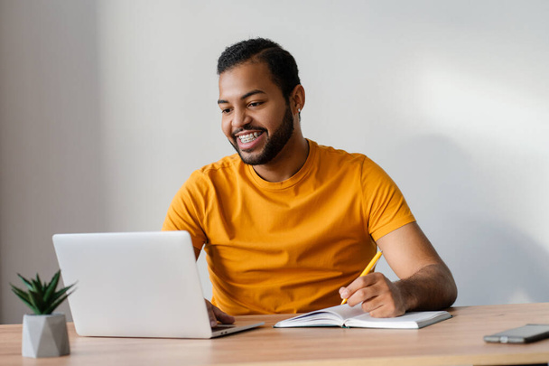 Hombre afroamericano sonriente con frenos dentales en los dientes usando computadora portátil estudiando, aprendiendo lenguaje. Tecnología, concepto de educación en línea  - Foto, imagen