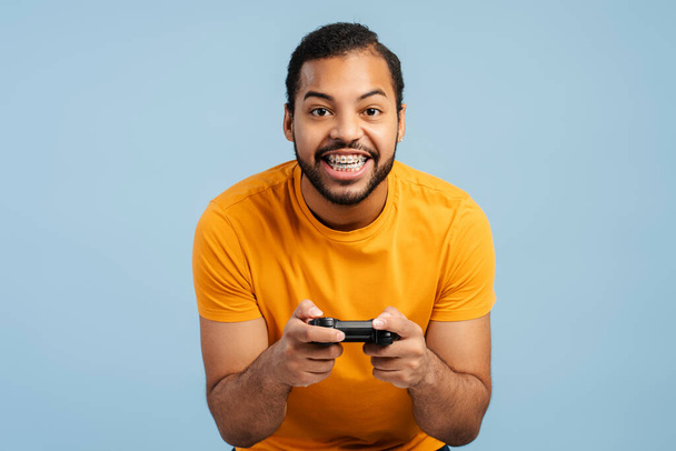 Emocionado hombre afroamericano con frenos, sosteniendo un controlador de juego y jugando, aislado en un fondo de estudio azul  - Foto, imagen
