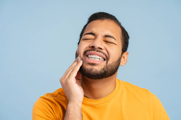 Portrait of troubled, african american man with braces, holding his cheek in pain, against a blue background. Illustrates dental discomfort concept  - Foto, immagini