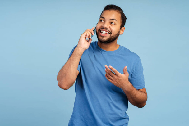 Glimlachende Afro-Amerikaanse man in gebitsbeugels praten tijdens mobiel bellen, zich gelukkig geïsoleerd voelen op een blauwe achtergrond  - Foto, afbeelding