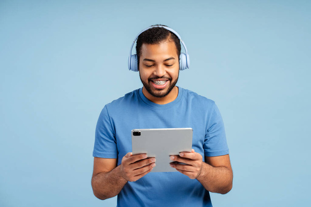 Ritratto di un bell'uomo sorridente che indossa le cuffie, tiene il tablet digitale, ascolta musica isolata su sfondo blu. Concetto divertente, tecnologia  - Foto, immagini