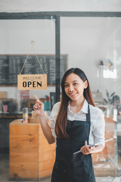 Portret van een vrolijke serveerster bij de ingang van het restaurant met open bord, Portret van een jonge zakenvrouw wonen nieuwe klanten in haar coffeeshop. - Foto, afbeelding