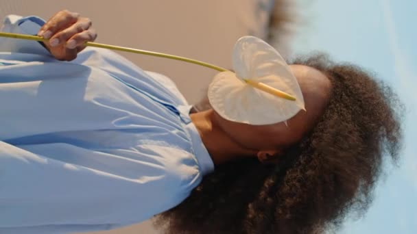 Ηρεμία ελκυστική κοπέλα που καλύπτει το πρόσωπο με λευκό λουλούδι στέκεται στην έρημο κάθετα closeup. Αφρο-Αμερικανίδα που ποζάρει κρατώντας την Κάλι, φοράει κομψό φόρεμα σε αμμώδεις αμμόλοφους. Κέρλι μοντέλο ψάχνει απόσταση - Πλάνα, βίντεο