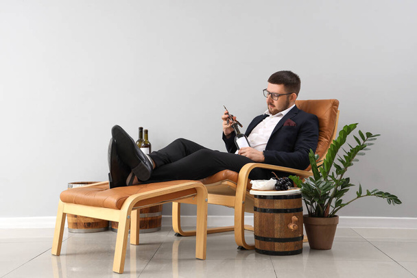 Молодой сомелье открывает бутылку вина, отдыхая на кресле с подставкой для ног - Фото, изображение