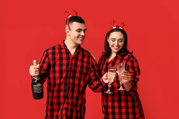 Счастливая молодая пара в рождественской пижаме и с шампанским на красном фоне - Фото, изображение