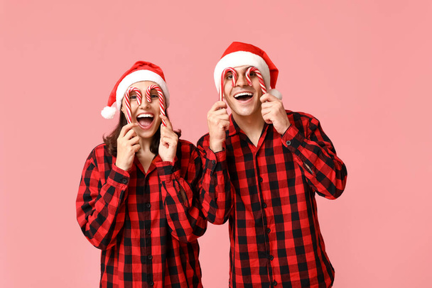 Funny young couple in Christmas pajamas and with candy canes on pink background - Photo, Image