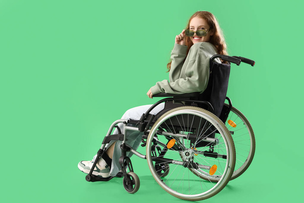 Yeşil arka planda güneş gözlüklü, tekerlekli sandalyedeki genç kızıl kadın. - Fotoğraf, Görsel