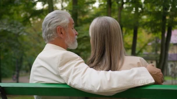 バックビューかわいいポジティブな70年代のカップルは,街の自然公園のベンチでリラックスして幸せな家族を引退しました. 既婚の配偶者コーカサス人男性女性は外で一緒に話す健康的なライフスタイルを楽しんで抱擁 - 映像、動画