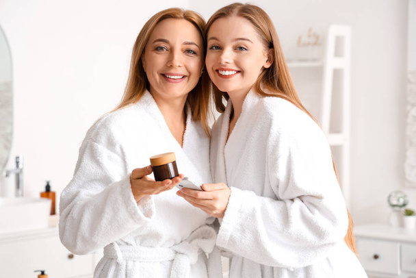Mujer joven y su madre con tarro de crema usando el teléfono móvil después de la ducha en el baño - Foto, imagen