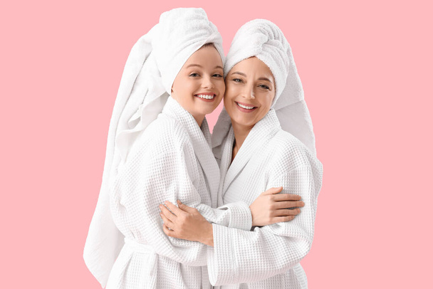 Mujer joven con su madre en albornoces después de la ducha abrazándose sobre fondo rosa - Foto, imagen