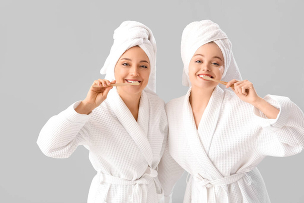 Young woman with her mother in bathrobes brushing teeth after shower on light background - Photo, Image