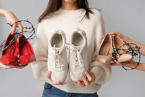 Jonge vrouw verkiest sneakers boven schoenen gewikkeld in prikkeldraad op grijze achtergrond. Oncomfortabel schoenen concept - Foto, afbeelding