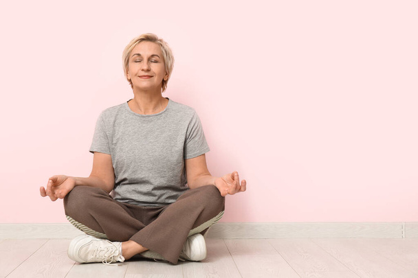 Portrait of meditating mature woman sitting on floor against pink wall - Photo, Image