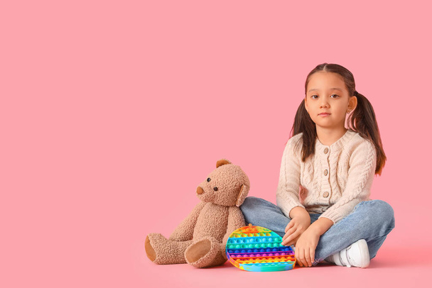 Klein Aziatisch meisje met beer en pop it fidget speelgoed op roze achtergrond. Autistische stoornis concept - Foto, afbeelding