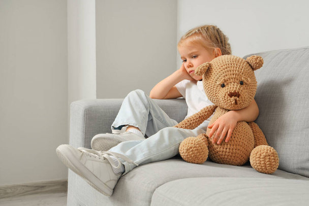 Грустная маленькая девочка с аутизмом и игрушечным медведем на диване дома - Фото, изображение