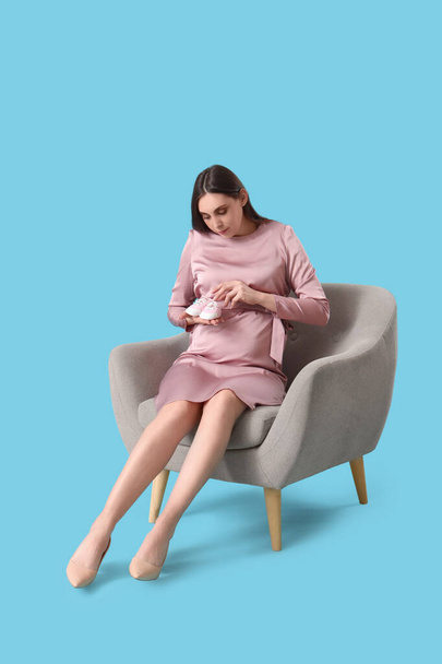 Young pregnant woman with baby booties sitting in armchair on blue background - Photo, Image