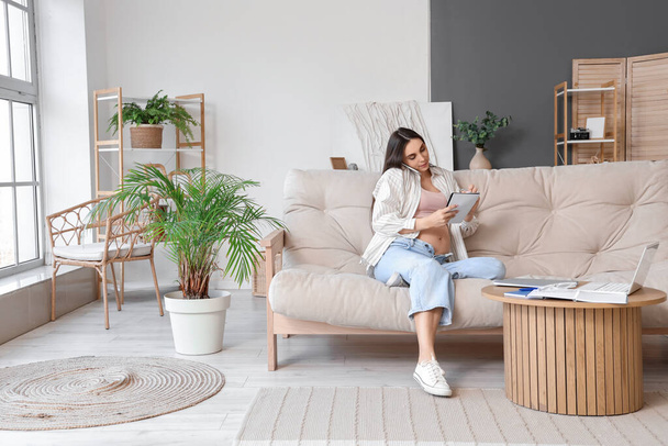 Mujer embarazada joven con portátil hablando por teléfono móvil en el sofá en casa - Foto, imagen
