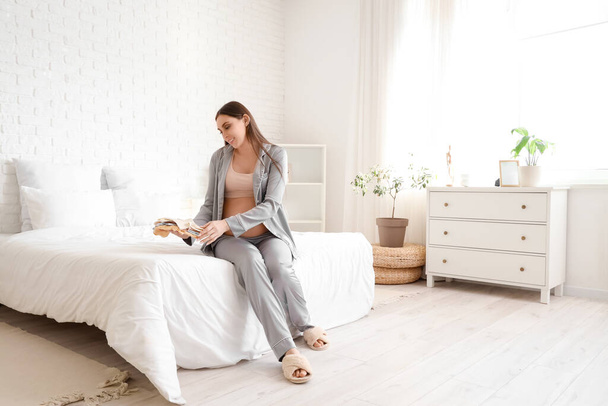 Jonge zwangere vrouw met baby bodysuits zitten in de slaapkamer - Foto, afbeelding