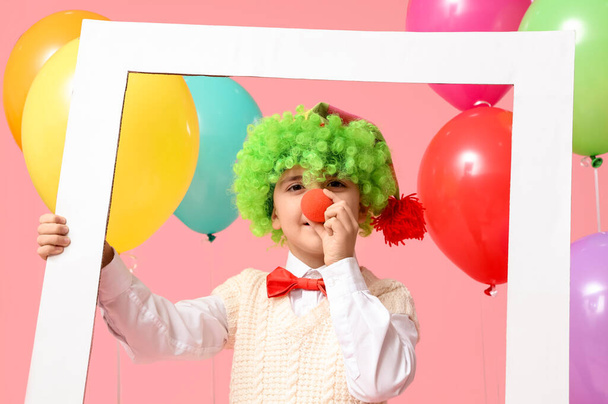 Słodki chłopczyk w peruce klauna i nosie z balonami trzymający ramkę na różowym tle. Święto prima aprilis - Zdjęcie, obraz