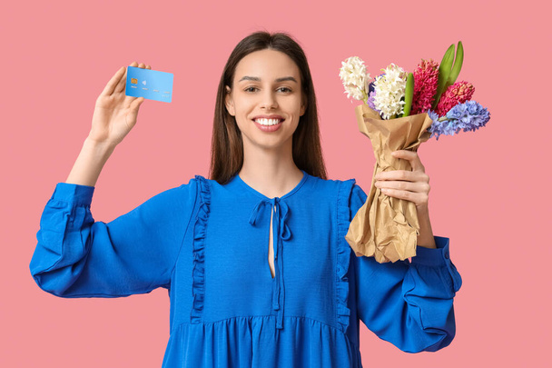 Glücklich lächelnde junge Frau mit Hyazinthenblüten und Kreditkarte auf rosa Hintergrund - Foto, Bild