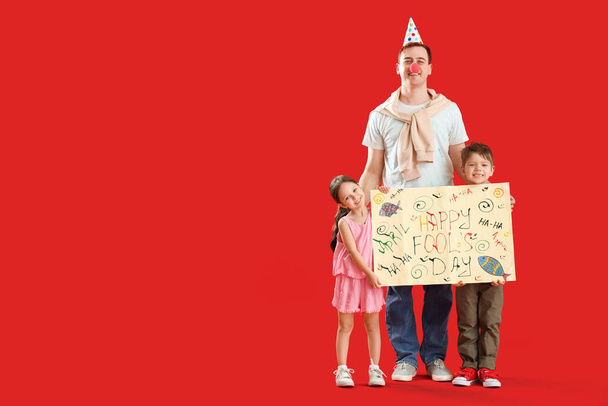 Ο πατέρας και τα μικρά παιδιά του κρατώντας αφίσα με κείμενο HAPPY FOOL 'S DAY σε κόκκινο φόντο - Φωτογραφία, εικόνα