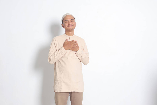 Beyaz gömlekli yakışıklı Asyalı Müslüman bir adamın portresi. Kafatası şapkası kalbine yerleştirilmiş, minnet duyuyor. Akıl sağlığı ve pozitif akıl kavramı. Beyaz arkaplanda izole edilmiş resim - Fotoğraf, Görsel