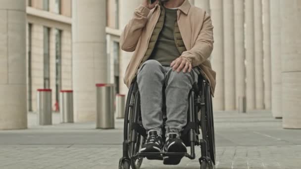 Naklonit záběr mladého podnikatele sedícího na invalidním vozíku a mající rozhovor na mobilním telefonu venku ve městě - Záběry, video