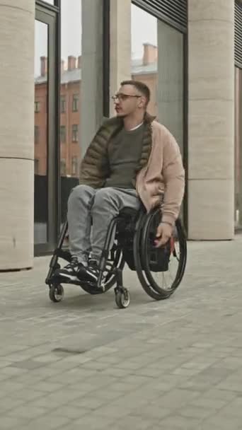 Colpo verticale di giovane in abiti casual cavalcare sedia a rotelle all'aperto lungo edificio urbano durante il giorno in città - Filmati, video
