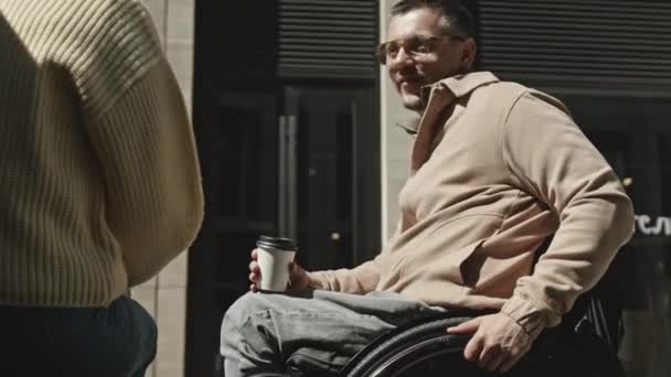 Tilt up shot of young man with disability sitting in wheelchair, drinking coffee from disposable cup and having talk with female friend outdoors on city street - Footage, Video