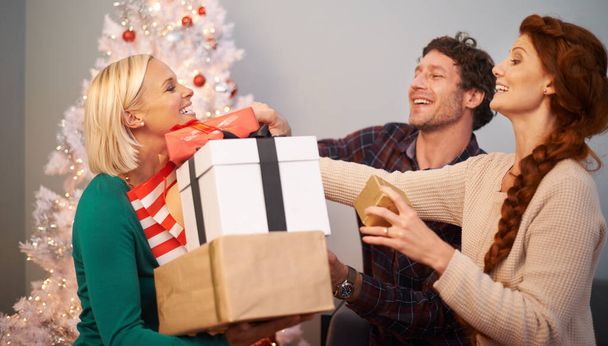 Φίλοι, Χριστούγεννα και δώρο για γιορτή στο δέντρο για εορταστική περίοδο ή δίνοντας κουτί δώρου, διακοπές ή διακοπές. Άνδρας, γυναίκες και αγροτεμάχιο στο σαλόνι για χειμερινές διακοπές ή ενθουσιασμένος, συγκόλληση ή αγάπη. - Φωτογραφία, εικόνα