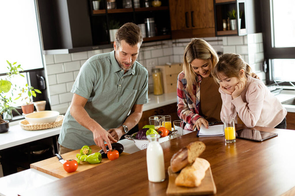 Junge Familie plaudert und bereitet Essen an einem geschäftigen Küchentisch mit frischen Zutaten und Kochutensilien zu - Foto, Bild