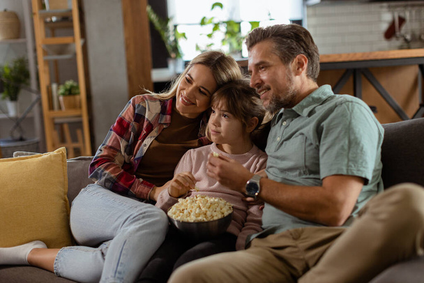 Una famiglia di tre persone è comodamente adagiata su un divano, i loro volti riflettono eccitazione e attenzione mentre condividono una ciotola di popcorn durante una serata cinematografica sospesa. - Foto, immagini