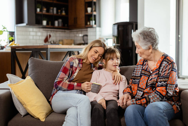 Trzy pokolenia kobiet śmieją się i rozmawiają na wygodnej kanapie w salonie - Zdjęcie, obraz